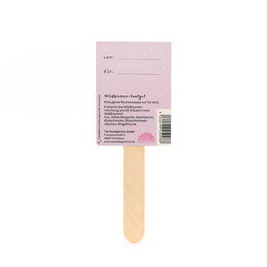Dos d'un sachet de graines de fleurs sauvages avec un bâton en bois et une étiquette violette indiquant les champs « de » et « pour » et une description du mélange de fleurs sauvages.