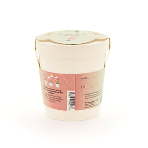 Pot de fleurs beige avec anse, étiquette rose avec illustrations de plantes et texte en allemand, étiquette cadeau sur le couvercle.
