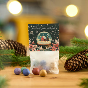 Sac avec des boules colorées et une carte de Noël avec l'inscription "Joyeux Noël" devant des décorations de Noël avec des pommes de pin et des branches.