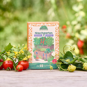 Un paquet de graines « Naschgarten » se trouve sur une table en bois dans le jardin, entouré de tomates fraîches, de fraises et de feuillage vert.