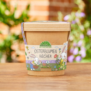 Tasse marron avec une banderole colorée représentant « Fleurs de Pùques dans une tasse » et deux lapins de Pùques dessinés avec des fleurs, debout sur une surface en bois dans le jardin.