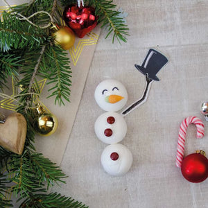 Un bonhomme de neige fait de guimauves avec un pain de sucre, des gouttes de chocolat et un nez de carotte repose sur un tissu à côté de décorations de Noël et de branches de sapin.