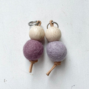 Deux porte-clés faits main avec des boules en bois et des boules en feutre de laine violettes sur fond blanc.