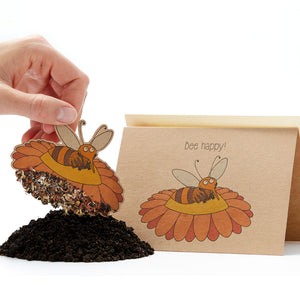 Une main tenant une carte de graines en forme d'abeille avec le slogan « Bee happy ! » dessus et un tas de terre en dessous.