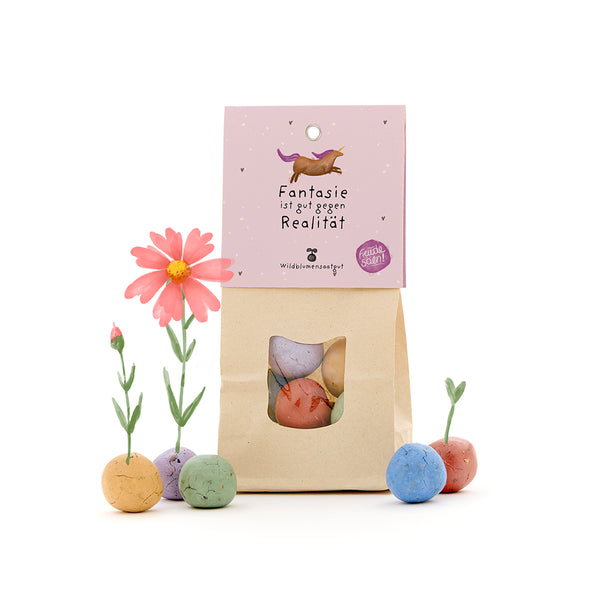 Sac en papier avec des boules de graines de fleurs sauvages colorées, certaines avec de petites fleurs, et une étiquette avec une licorne et le texte « L'imagination est bonne contre la réalité ».