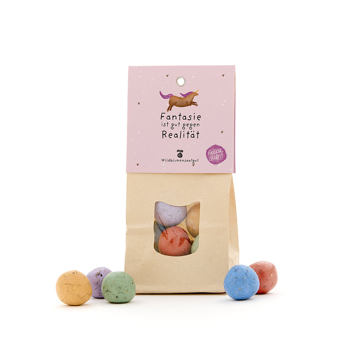 Sac en papier avec boules de graines colorées, étiquette avec licorne et texte « L'imagination est bonne contre la réalité. »