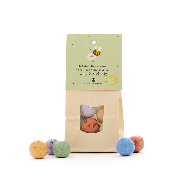 Un sac en papier brun avec une fenêtre, rempli de boules de graines colorées et une étiquette verte avec une illustration d'abeille et un dicton allemand.