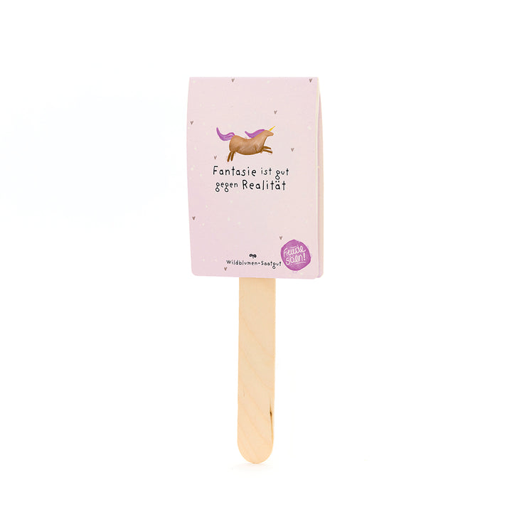 Bâton en bois avec une carte rose, dessus une licorne dessinée et le texte « L'imagination est bonne contre la réalité », en dessous duquel il est écrit « Graines de fleurs sauvages ».