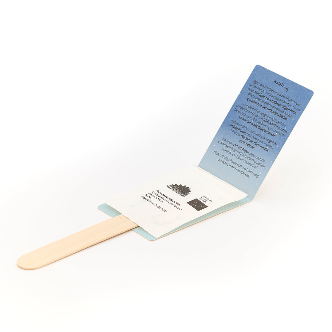 Bâtonnet de glace en bois avec sachet de graines attaché et instructions dépliées en allemand.