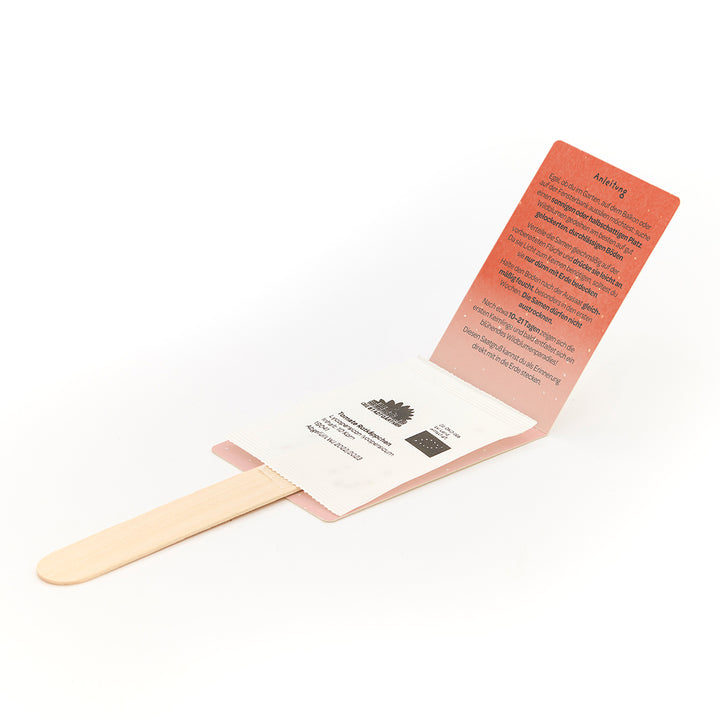 Bâton en bois avec sachet de graines attaché et instructions dépliées en allemand.