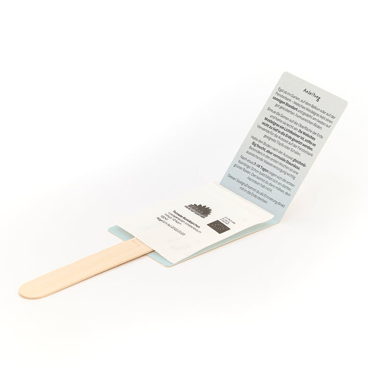 Un bâton en bois avec un emballage ouvert contenant un sachet de graines et des instructions imprimées en allemand.