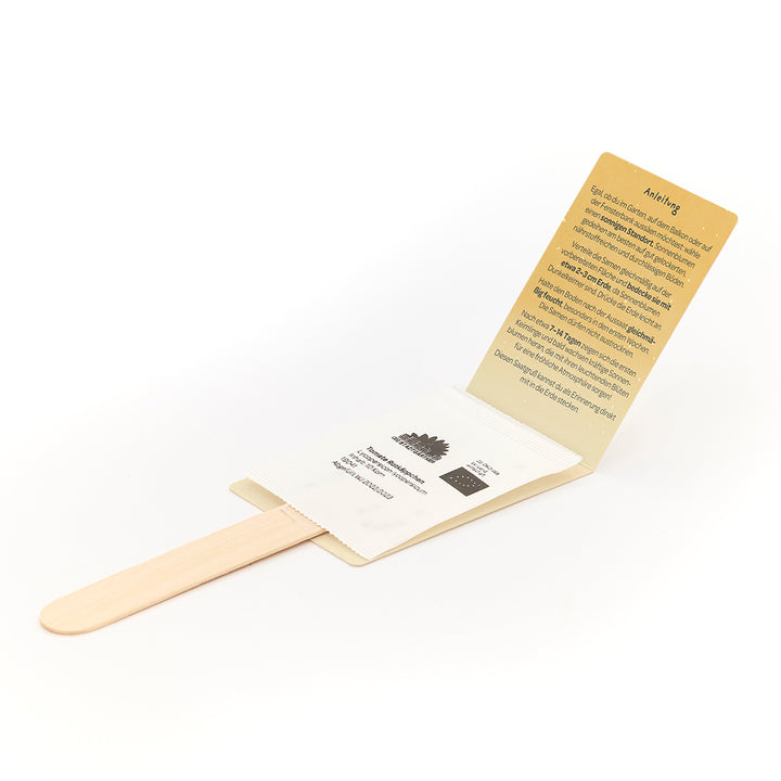 Bâton en bois avec enveloppe attachée contenant des graines et instructions dépliantes en allemand.