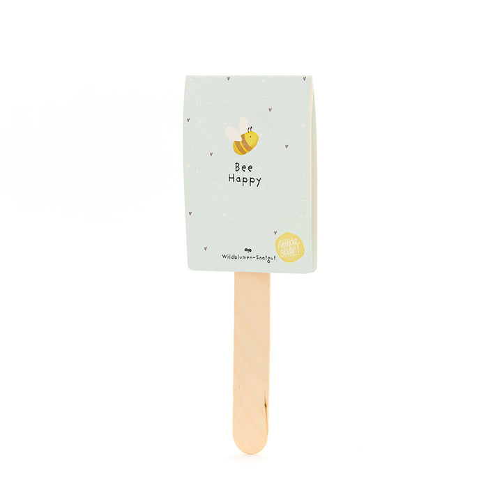 Bâton en bois avec une étiquette bleu clair comportant un dessin d'abeille et le texte "Bee Happy" ; graines de fleurs sauvages.