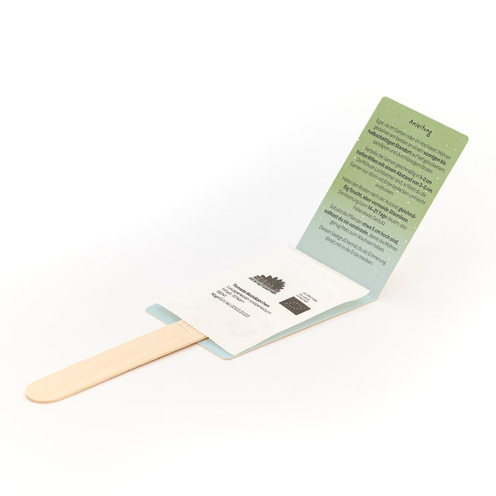 Un ensemble pliable avec un bâton en bois et un sachet de graines, ainsi qu'une carte d'instructions verte en allemand.