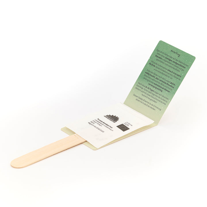 Bâton en bois avec enveloppe attachée contenant une carte d'instructions verte et un sachet de graines blanches.