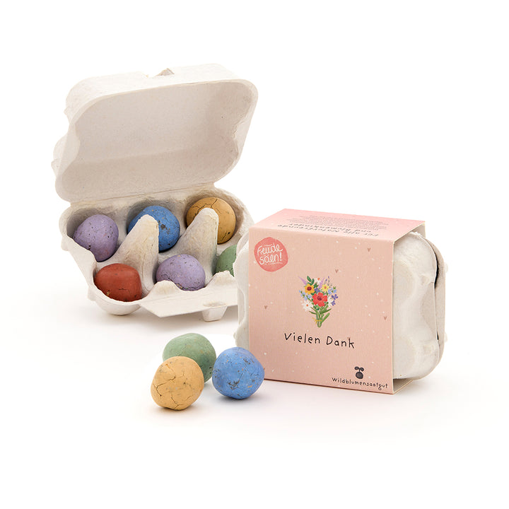 Six bombes de graines colorées dans un petit paquet en carton d'œufs, à côté d'une boîte fermée avec l'inscription « Merci » et une illustration de fleur.