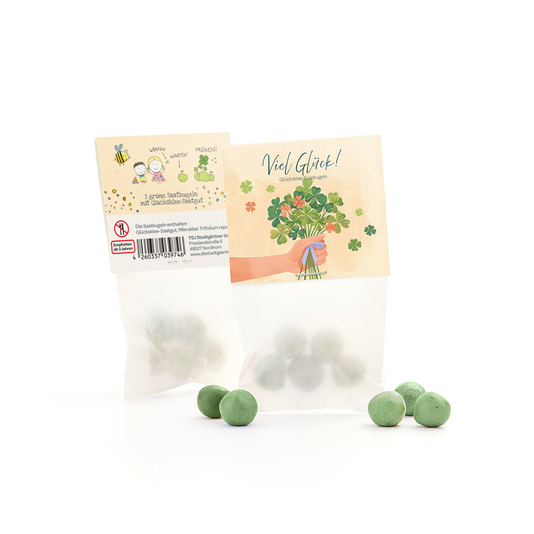 Sachet avec boules de graines vertes et graines de trèfle porte-bonheur, à côté de plusieurs boules vertes en vrac, sur l'emballage un motif de félicitations et une illustration de trèfle.