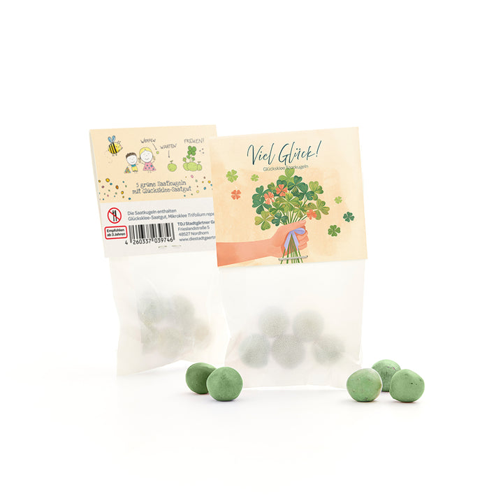 Sachet avec boules de graines vertes et graines de trèfle porte-bonheur, à côté de plusieurs boules vertes en vrac, sur l'emballage un motif de félicitations et une illustration de trèfle.