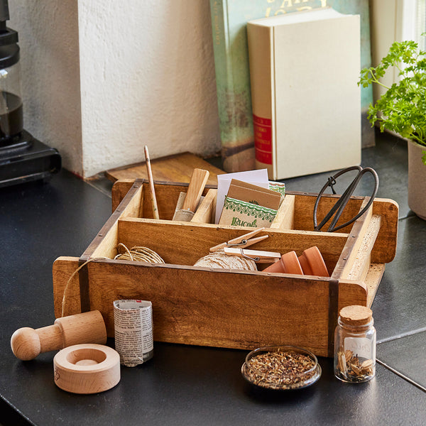 Boîte en bois contenant des outils de jardinage, notamment des sachets de graines, une pelle, des ciseaux, des petits pots en argile, de la ficelle et un pot à herbes, sur un comptoir de cuisine.