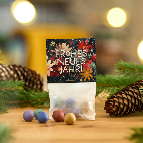 Un sac de boules colorées et une carte sur laquelle est écrit « Bonne année ! » reposent sur une table en bois, entourée de branches de sapin et de pommes de pin.