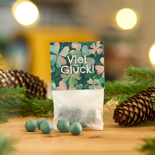 Sacs avec des boules vertes et l'inscription "Bonne chance !" devant des décorations de Noël avec des branches de sapin et des pommes de pin.