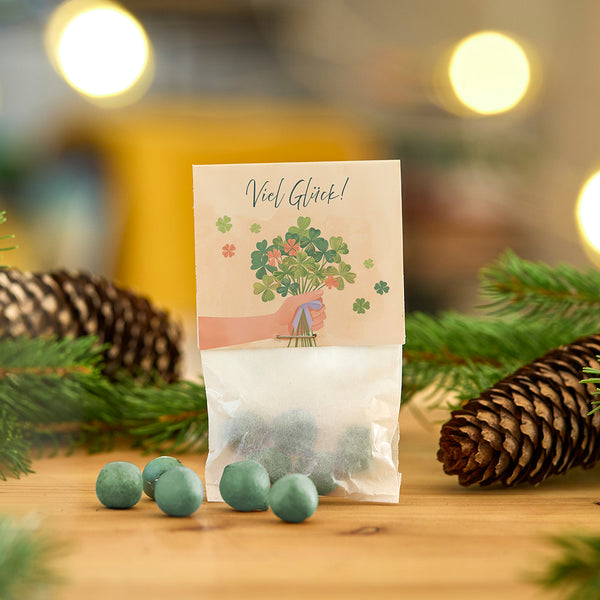 Sac de boules verdâtres, décoré d'une carte avec l'inscription « Bonne chance ! » et d'un bouquet de trèfles entouré de branches de sapin et de cônes.
