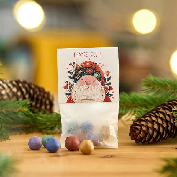 Sac avec boules colorées, carte de Noël avec motif du Père Noël et l'inscription « Joyeux Noël ! », pommes de pin et branches en arrière-plan.
