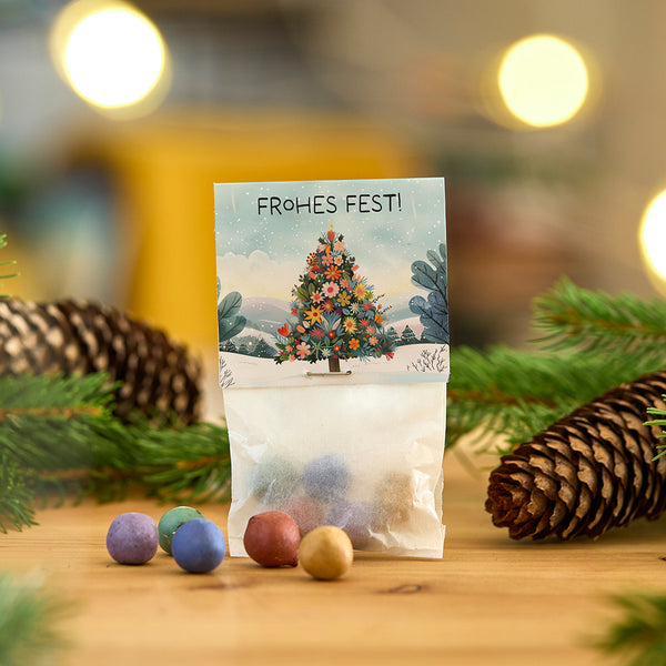Sac de boules colorées devant une carte avec un sapin de Noël et les mots « Joyeux Noël », entouré de branches de sapin et d'une pomme de pin.