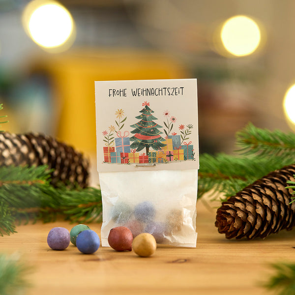 Un petit sac de boules colorées, décoré d'une carte de Noël avec l'inscription « Joyeux Noël », entourée de branches de sapin et d'une pomme de pin.