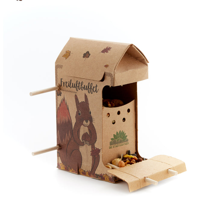 Mangeoire en carton avec motif écureuil et inscription "Buffet en plein air", remplie de noix et de graines.