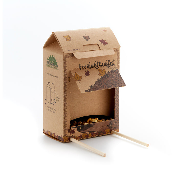 Boîte en carton avec rabat ouvert, feuilles d'automne, illustration de hérisson et deux bâtons en bois, sert de station d'alimentation pour les hérissons.