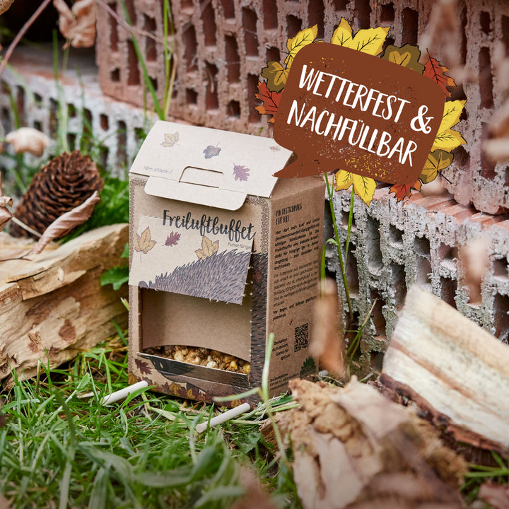Un carton étiqueté « Buffet en plein air » servant de mangeoire pour animaux résistante aux intempéries et rechargeable se trouve dans l'herbe à côté de bois et de briques.