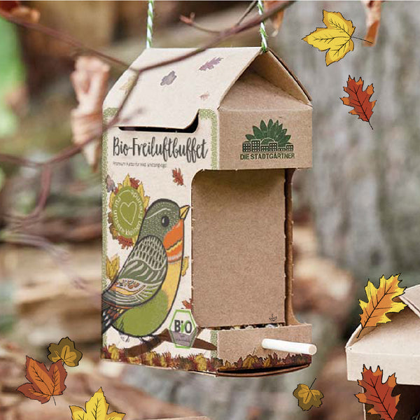 Mangeoire à oiseaux en carton avec des feuilles d'automne, un motif d'oiseau coloré et l'inscription « Buffet bio en plein air » suspendue à une branche.