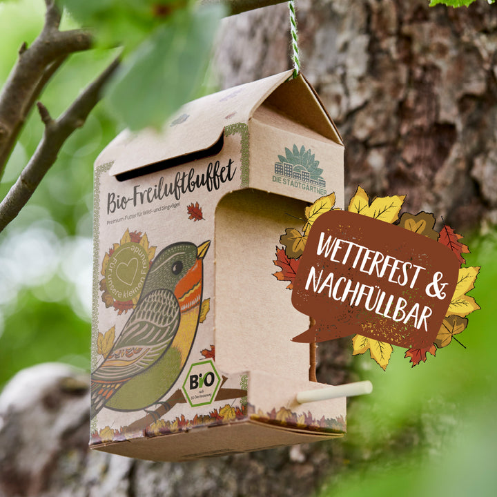 Une mangeoire à oiseaux en carton avec une illustration d'oiseau et l'inscription « Buffet bio en plein air » est accrochée à un arbre, à côté d'elle le texte « Résistant aux intempéries et rechargeable ».