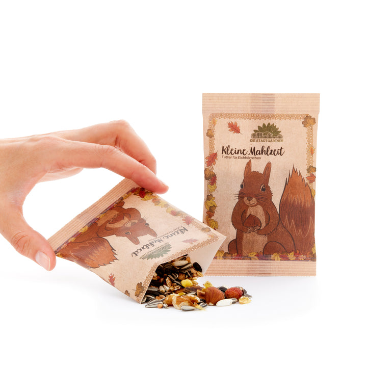 La main tient un sac en papier contenant de la nourriture pour écureuil, d'où tombent des graines et des noix sur une surface blanche, à côté d'un autre sac avec une illustration d'écureuil.