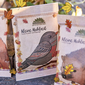 Sac en papier avec un imprimé d'oiseau et l'inscription "Small Meal - Food for Wild and Songbirds" de Die Stadtgärtner, entouré de feuilles d'automne.