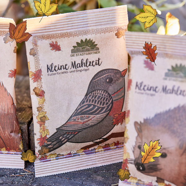 Sac en papier avec un imprimé d'oiseau et l'inscription "Small Meal - Food for Wild and Songbirds" de Die Stadtgärtner, entouré de feuilles d'automne.