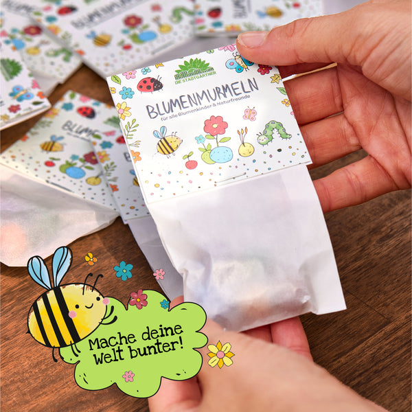 Une main tient un sachet de graines colorées étiqueté « Flower Marbles » au-dessus d'une table en bois ; au premier plan se trouve un dessin d'une abeille avec le texte « Rendez votre monde plus coloré ! »