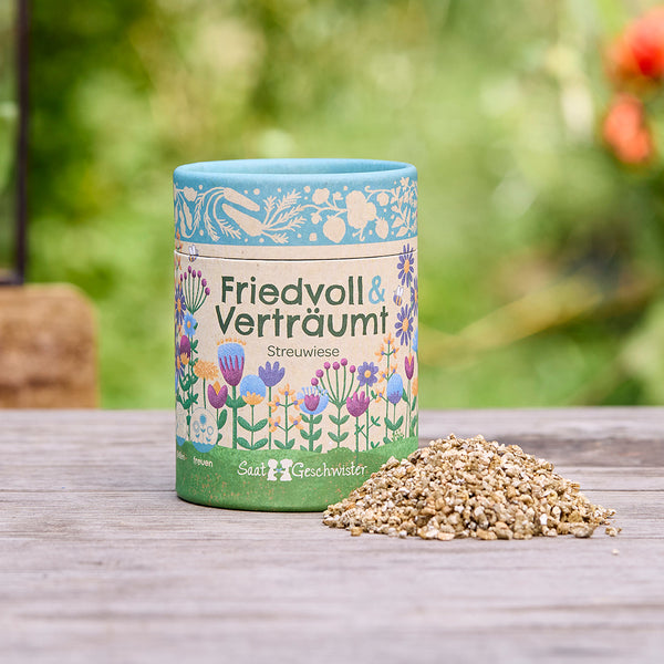 Boîte de graines colorée avec l'inscription « Peaceful & Dreamy Scattered Meadow » et un tas de graines de fleurs sur une table en bois extérieure.