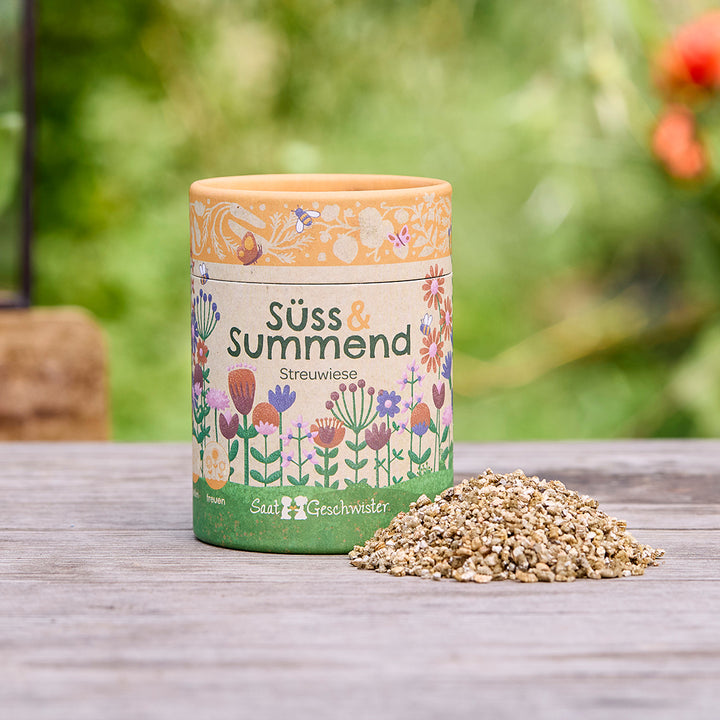Boîte ronde avec l'inscription « Sweet & Buzzing Meadow » et un motif floral, à côté un petit tas de graines sur une table en bois, fond vert flou.
