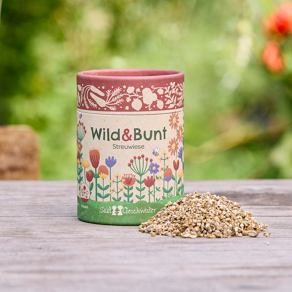 Une boîte avec l'inscription « Wild & Bunt Streuwiese » et des motifs floraux illustratifs, à côté d'un petit tas de graines, sur une table en bois à l'extérieur.