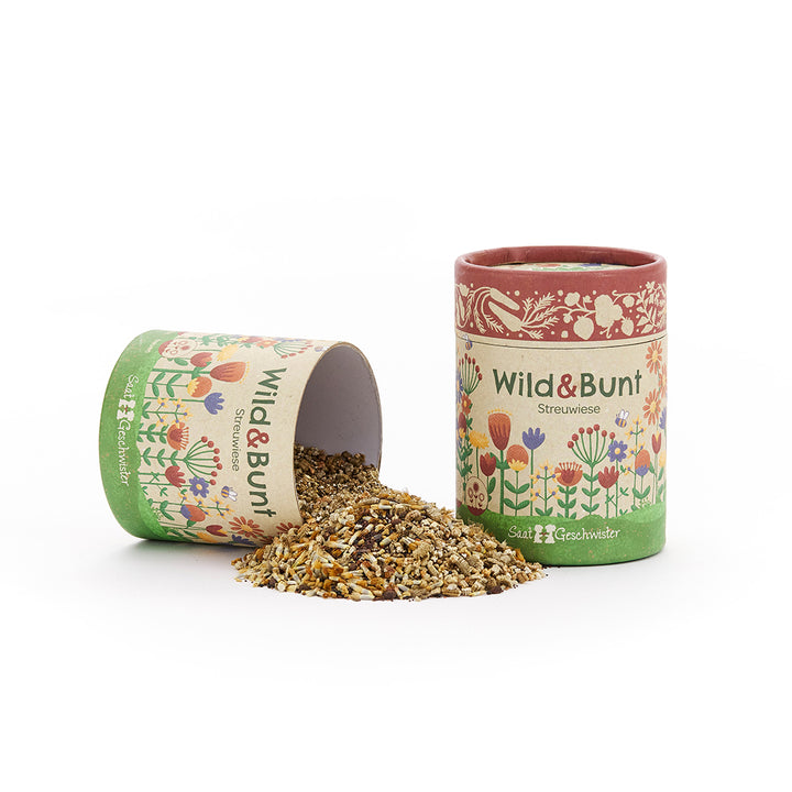 Deux boîtes colorées avec l'inscription « Wild & Colorful Streuwiese » (Dispersion de prairies sauvages et colorées), l'une couchée et ouverte, d'où tombent les graines sur la table.
