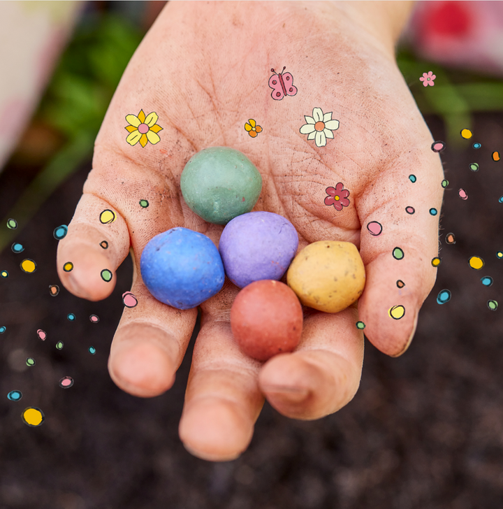 Une main tient cinq bombes de graines colorées, entourées de fleurs dessinées, de points et d'un papillon.
