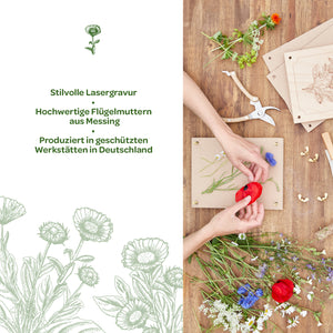 Deux mains disposent des fleurs colorées sur un cadre en bois ; des outils, des écrous papillon en laiton et des plaques de bois gravées sont posés à côté, sur une table en bois. À gauche, un texte présentant des informations sur la gravure laser élégante et la production en Allemagne.
