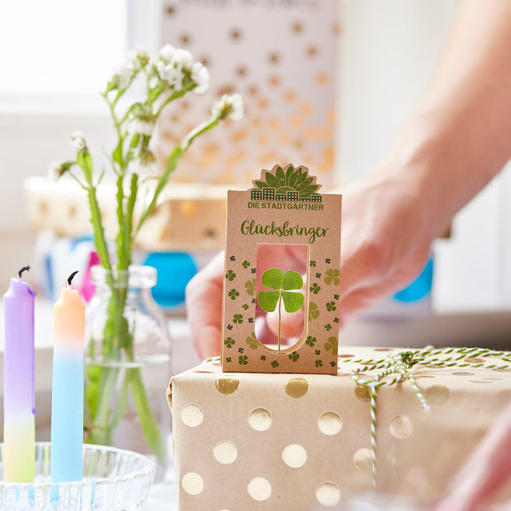 Une main tient un cadeau emballé avec un trèfle à quatre feuilles et les mots « Lucky Charm » au-dessus, avec des fleurs et des bougies colorées visibles en arrière-plan.