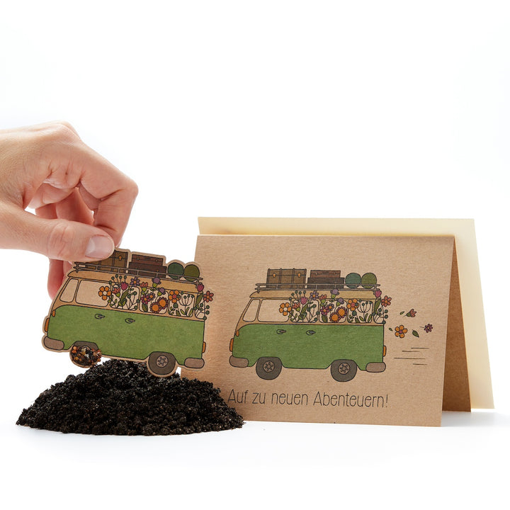 La main plante dans le sol une carte verte en forme de camionnette avec un motif de fleur, à côté une carte de vœux avec le texte « En route vers de nouvelles aventures ! ».