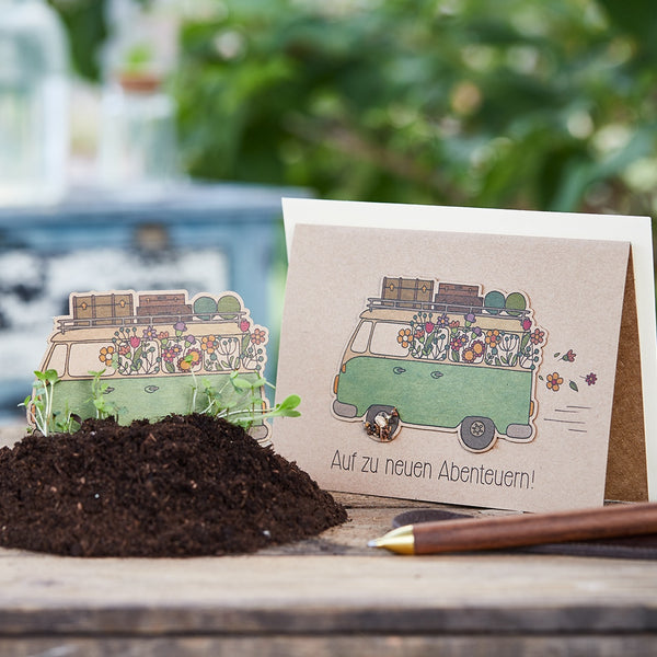 Carte de vœux avec un bus vert rempli de fleurs et de bagages, à côté de la terre avec de petites plantes, texte sur la carte : "En route vers de nouvelles aventures !"