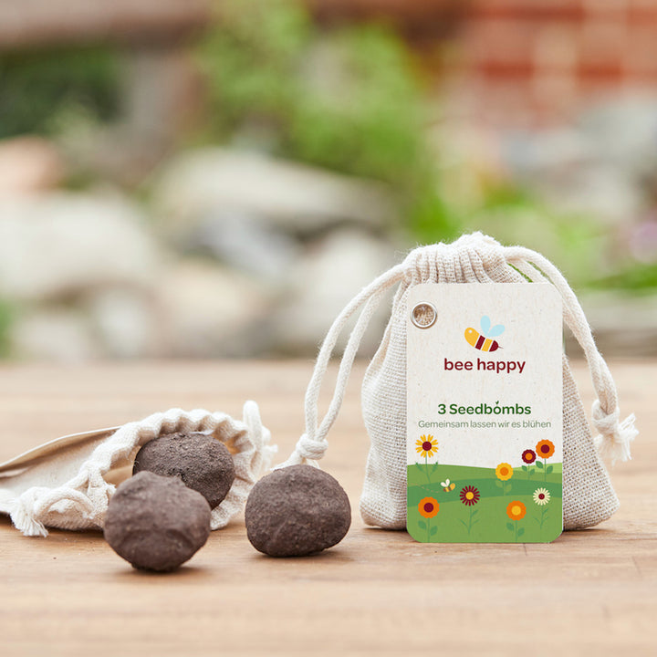 Des bombes à graines à côté d'un sac étiqueté « bee happy 3 seed bombs » sur une table en bois avec un fond vert flou.