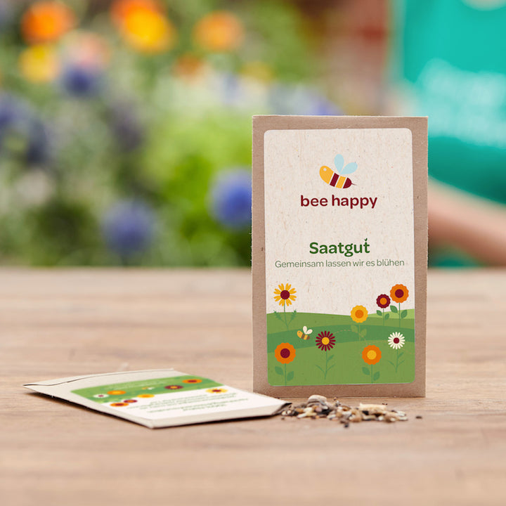 Sac de graines de fleurs « Bee Happy » sur une table en bois, avec des fleurs floues en arrière-plan.