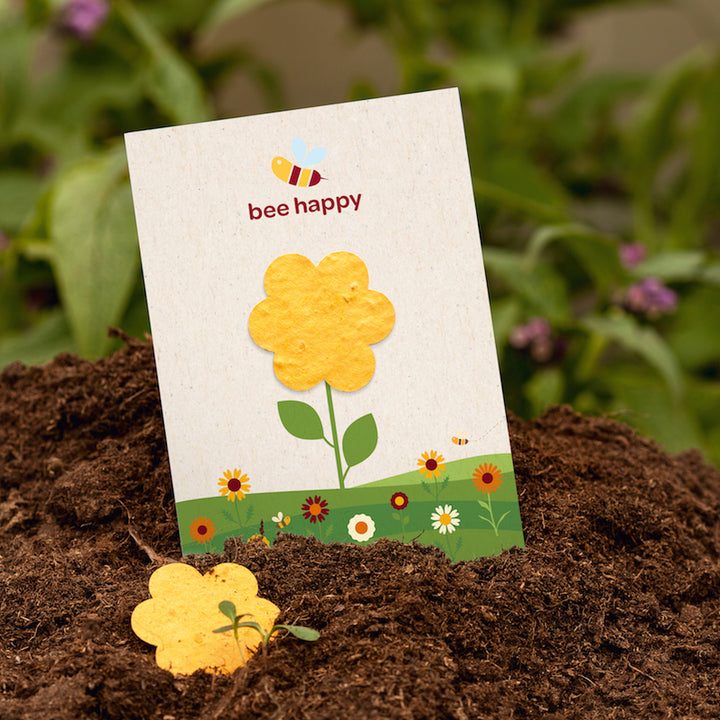Une carte avec une fleur jaune et l'inscription « abeille heureuse » est collée dans le sol ; au premier plan se trouve une fleur en papier jaune ; à l'arrière-plan se trouvent des plantes vertes.